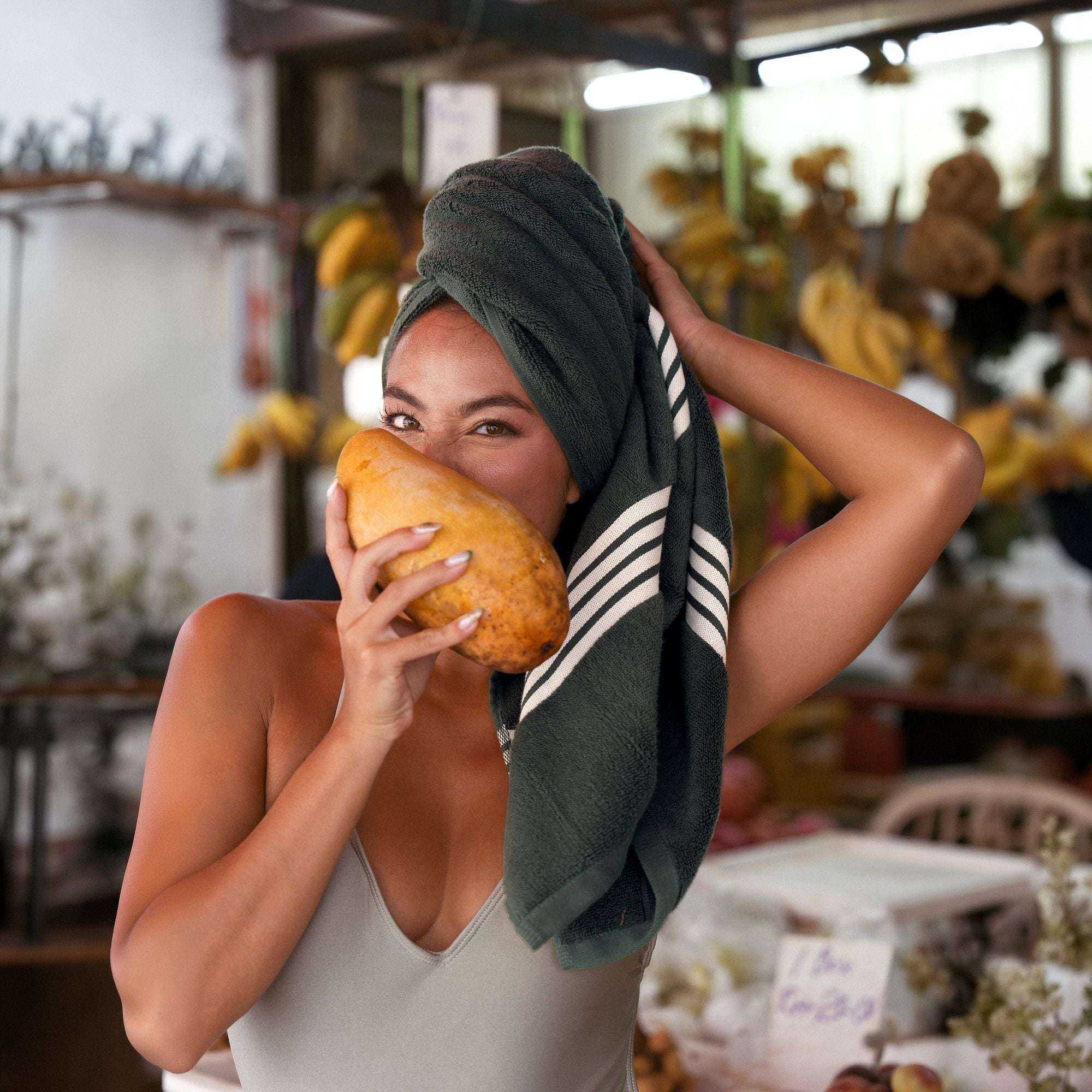Australian Cotton towels in dark green with white geometric stripes wrapped on a woman holding a yellow papaya, showcasing softness and absorbency
