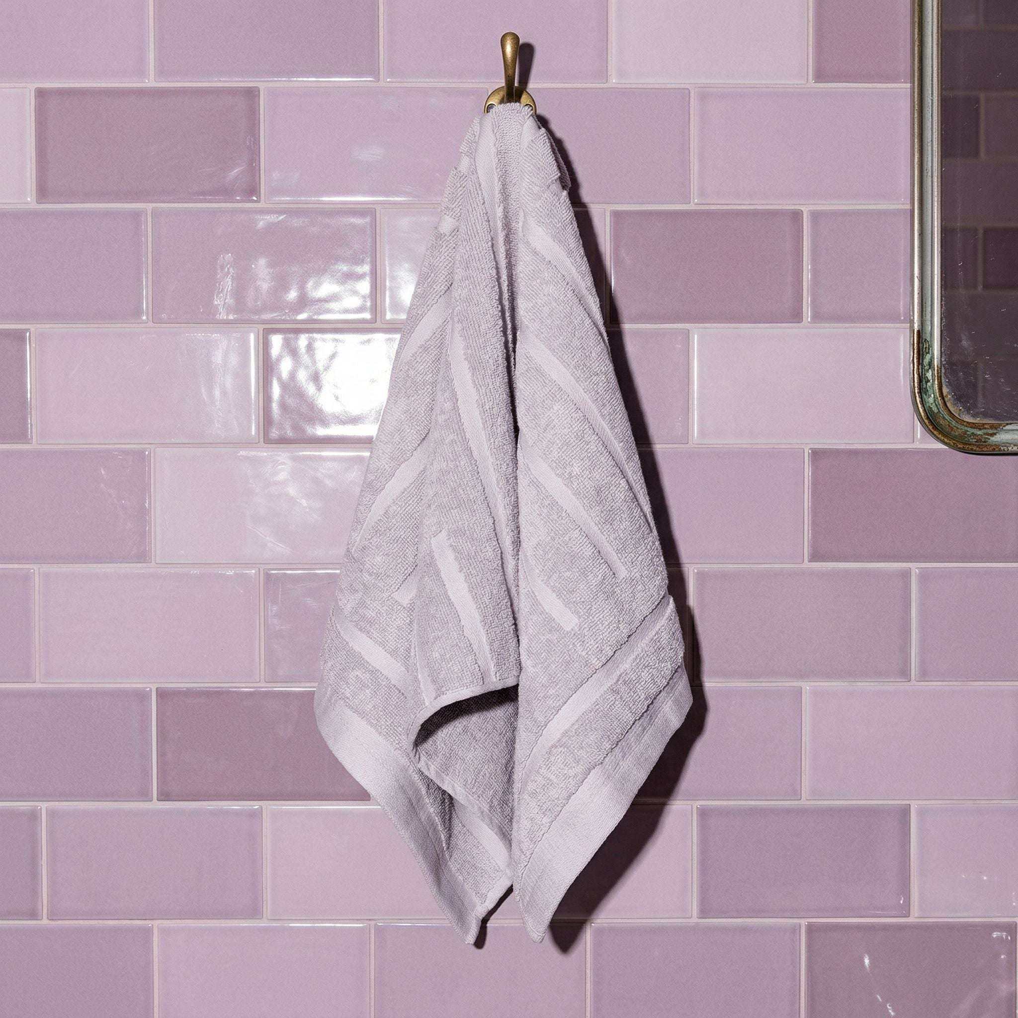 Australian Cotton towels in lilac color with geometric patterns hanging on a gold hook against pastel purple tiled wall