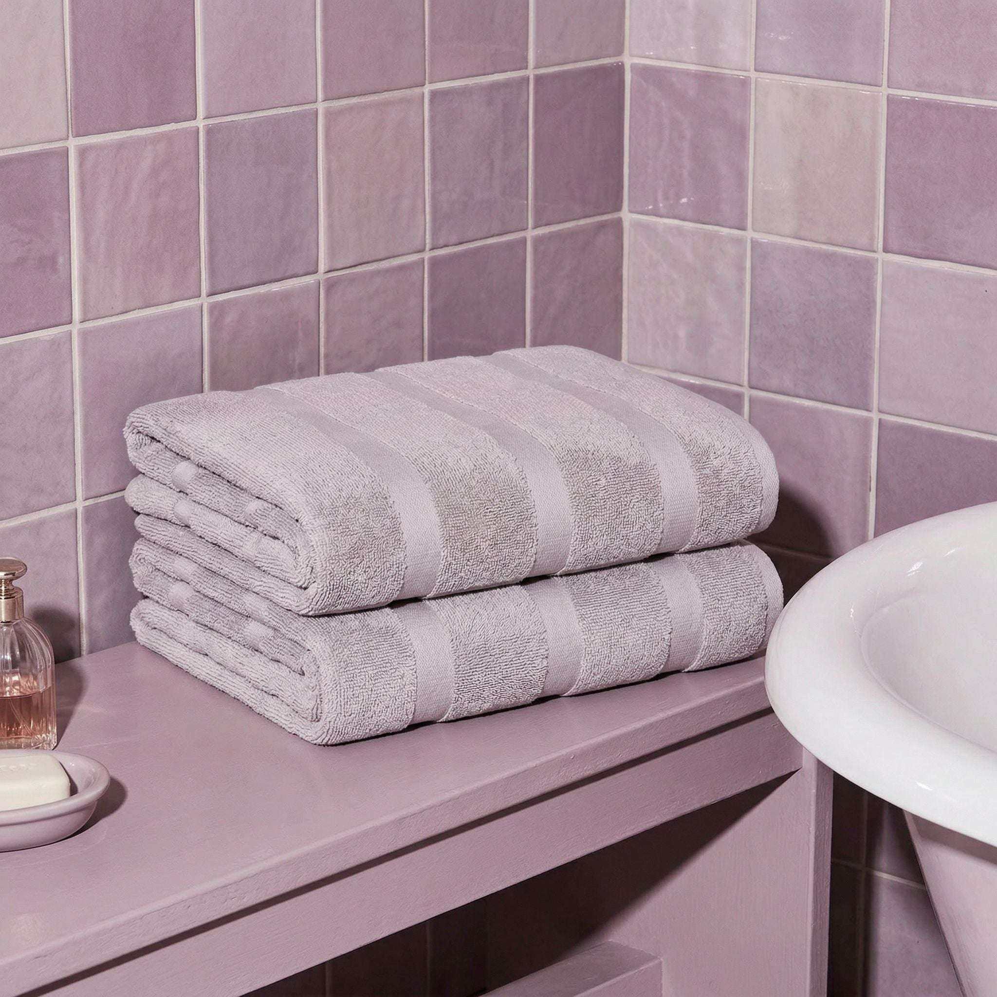 Australian Cotton towels in soft lilac color with subtle striped pattern folded next to white basin on purple tiled surface
