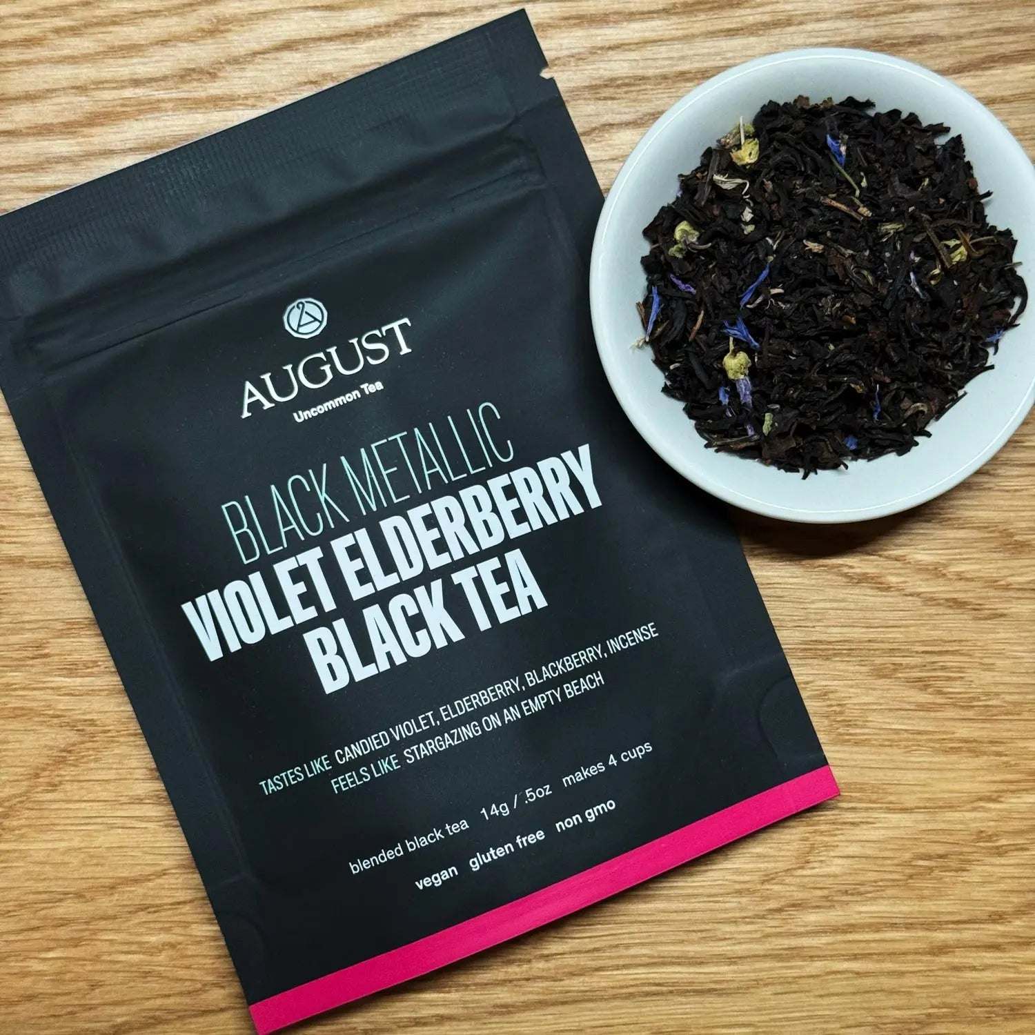 Black Metallic - Violet Elderberry Black Tea blend in a matte black pouch with a pink stripe and loose dark leaves with blue petals