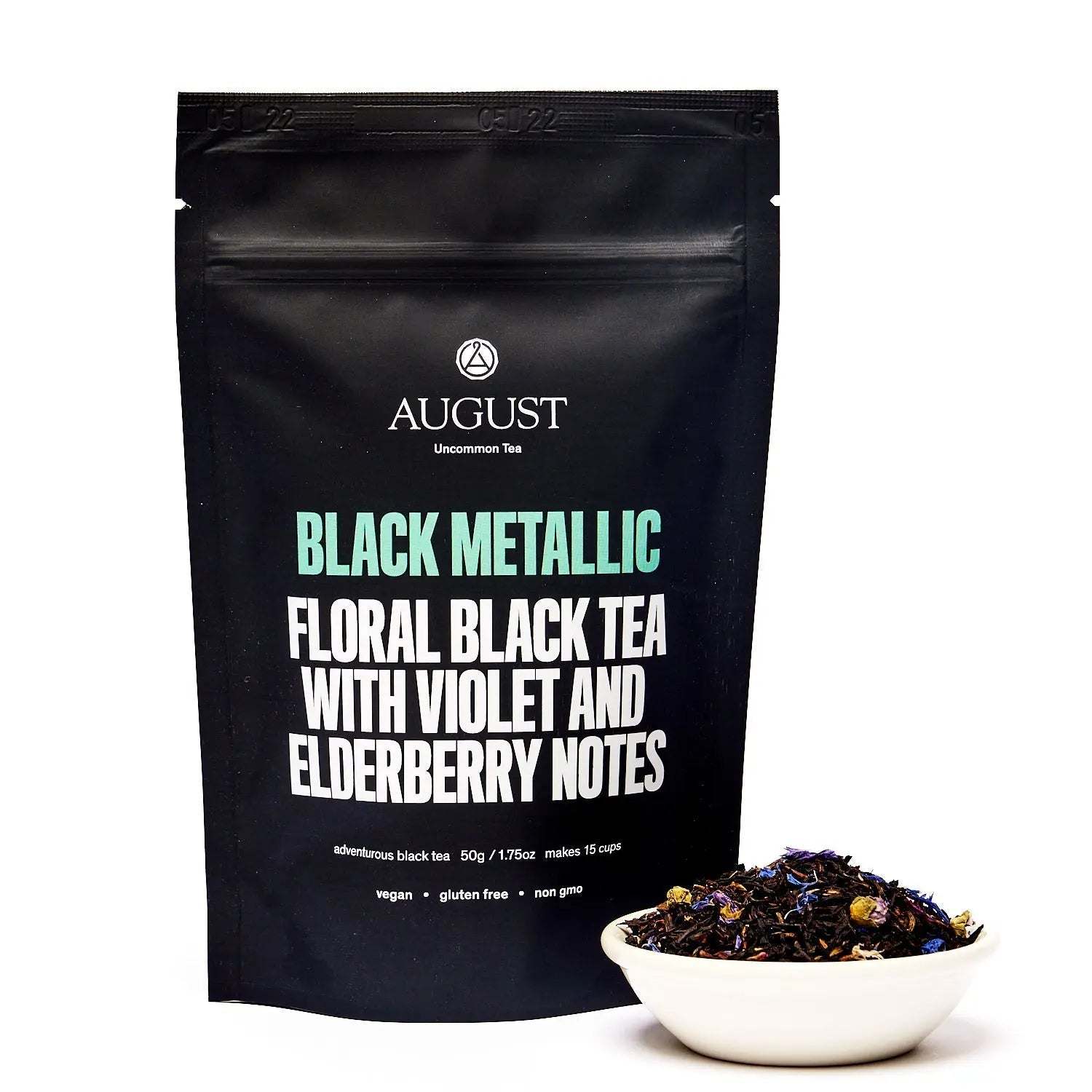 Black tea pouch in matte black packaging with teal and white lettering beside a bowl of dark loose tea leaves.