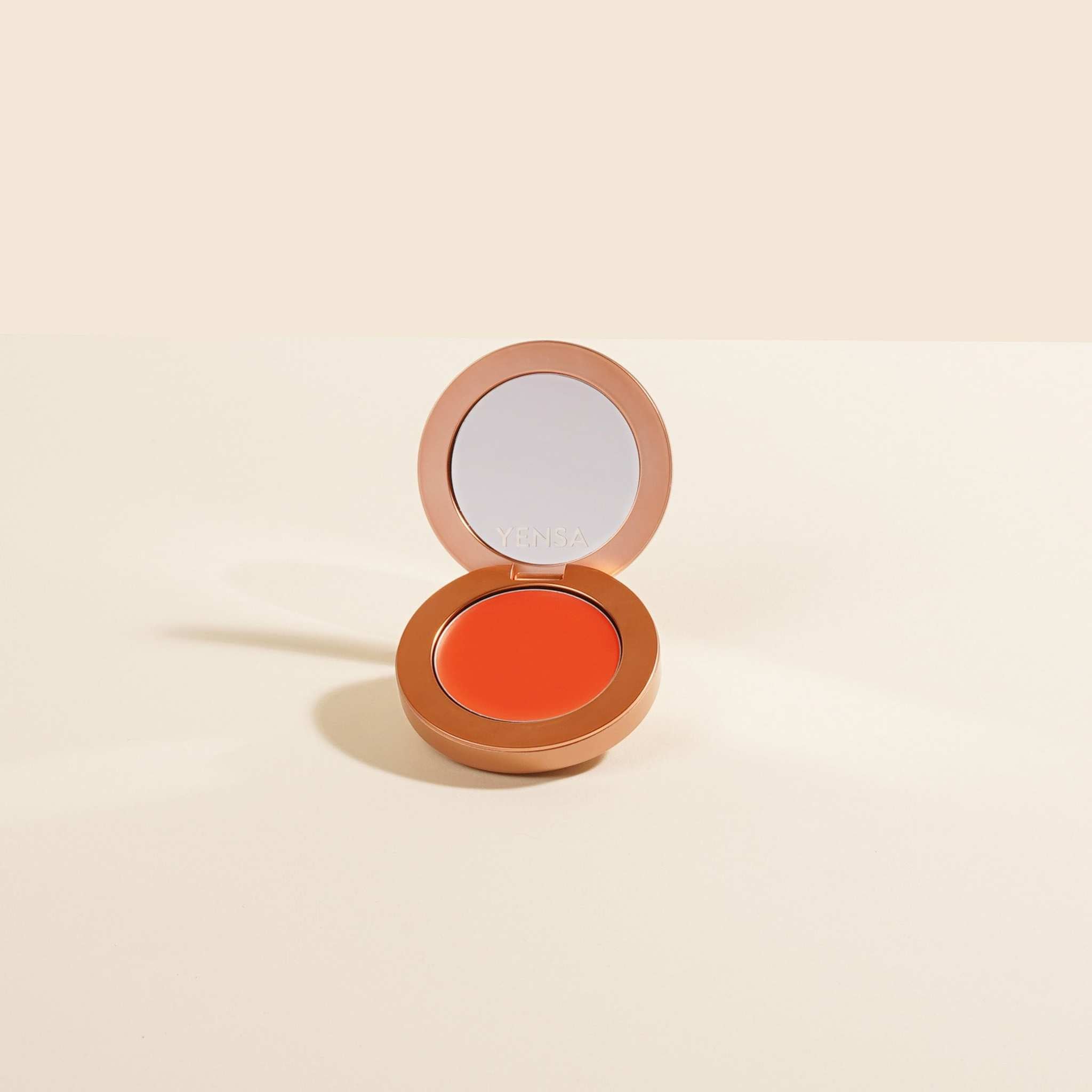 Buildable Cream Blush in a warm coral-orange cream formula, housed in a round copper compact with mirror lid