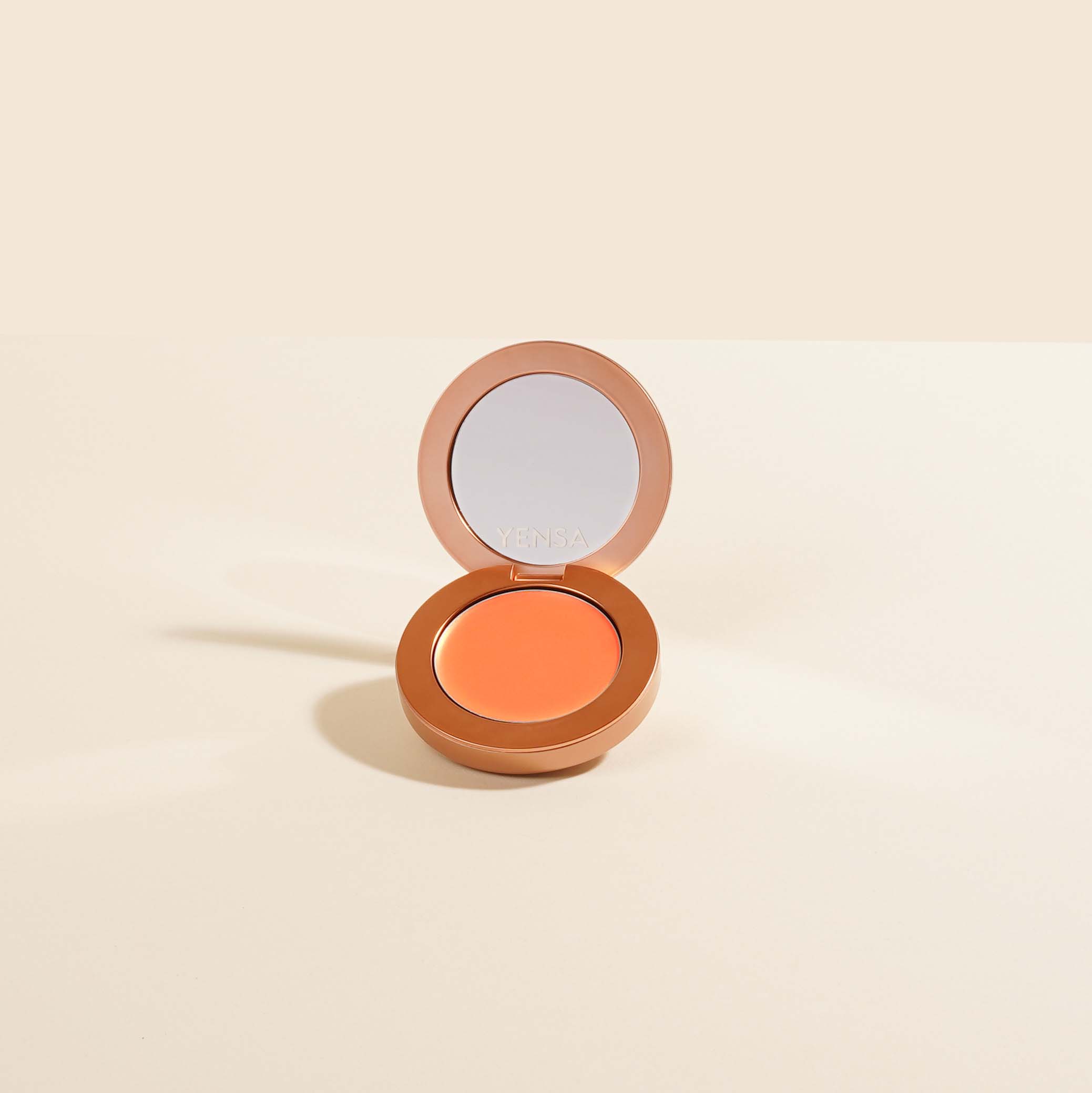Buildable Cream Blush in a warm coral shade, housed in a round metallic bronze compact with mirror lid