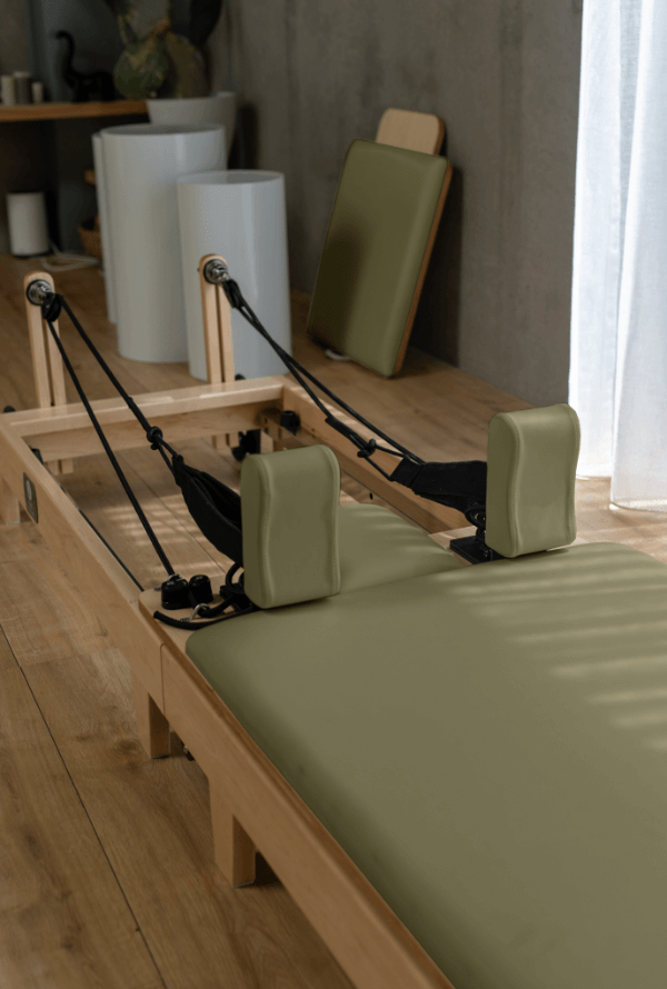 Eco Folding Pilates Reformer with a light wood frame and olive green padded carriage, shoulder rests and headrest.