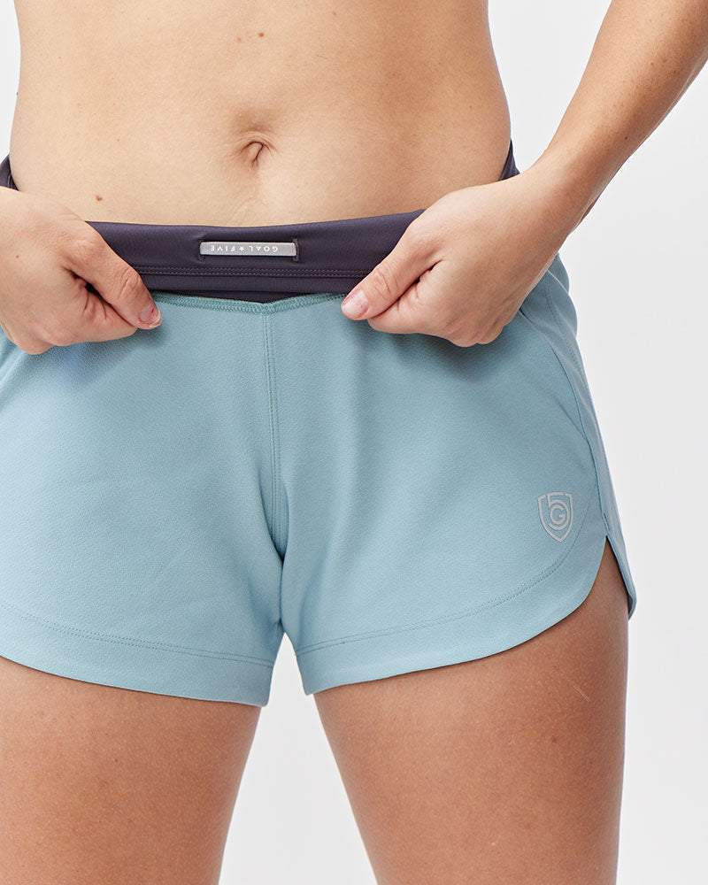 Excel Women's Workout Shorts in light blue stretch fabric with a dark waistband, small shield logo, and side slits