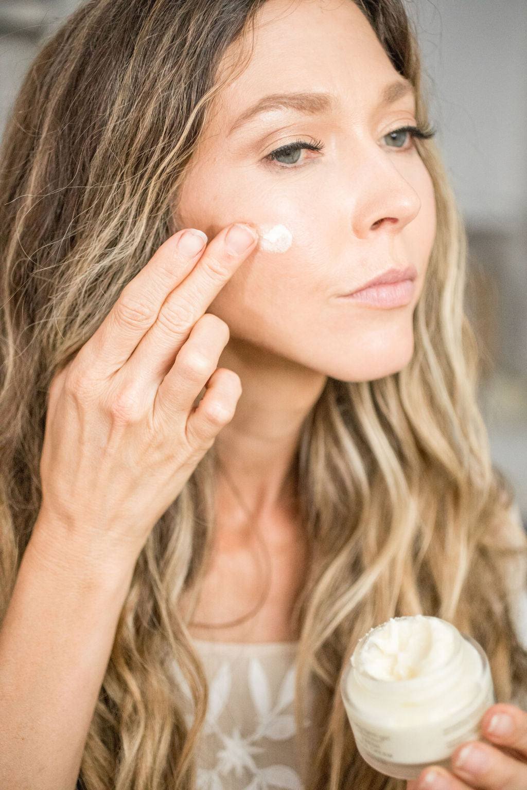 Fresh Face Cream, a woman dabs white moisturizer onto her cheek while holding an open glass jar with a black lid