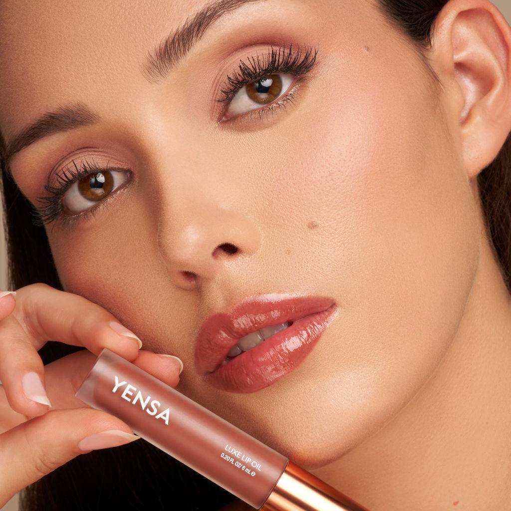 Luxe Lip Nourishing Oil in a rose-brown shade, shown in a clear tube with a copper-toned cap beside glossy lips