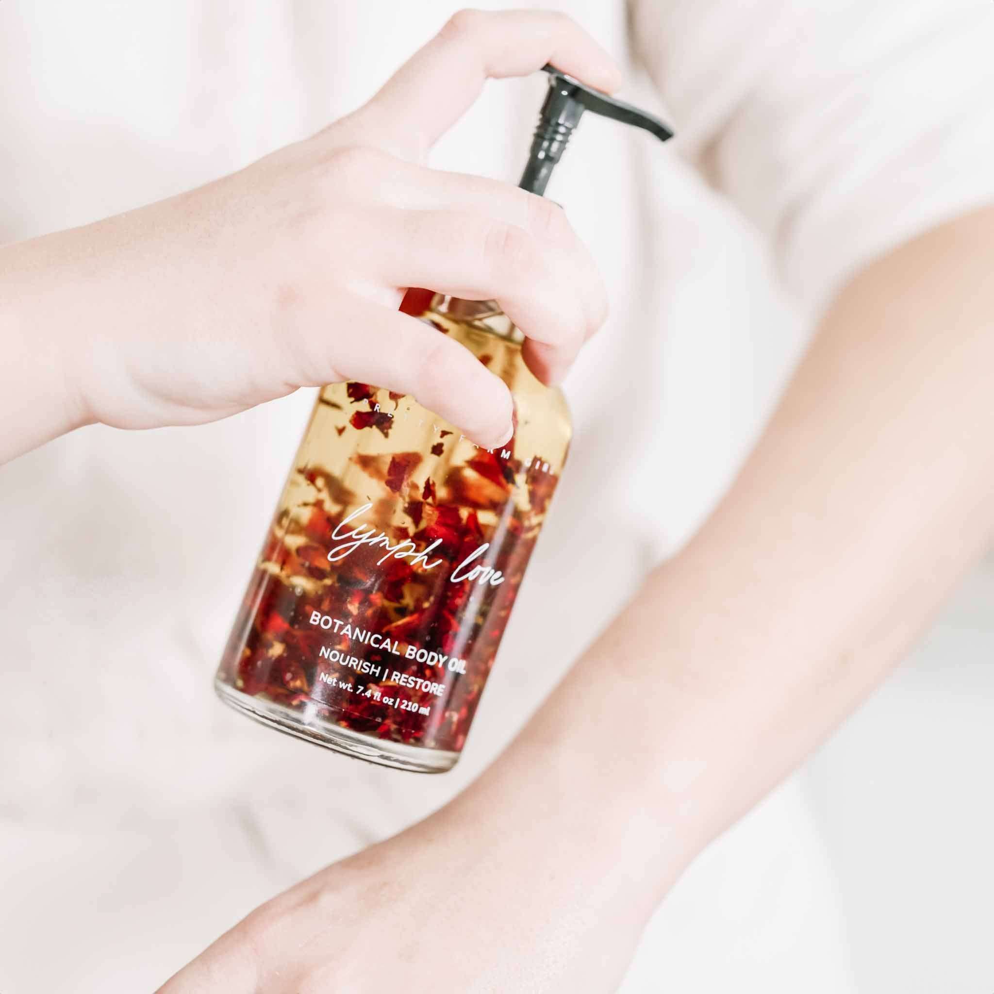 Lymph Love Body Massage Oil in a clear glass pump bottle with amber oil and visible red botanical petals