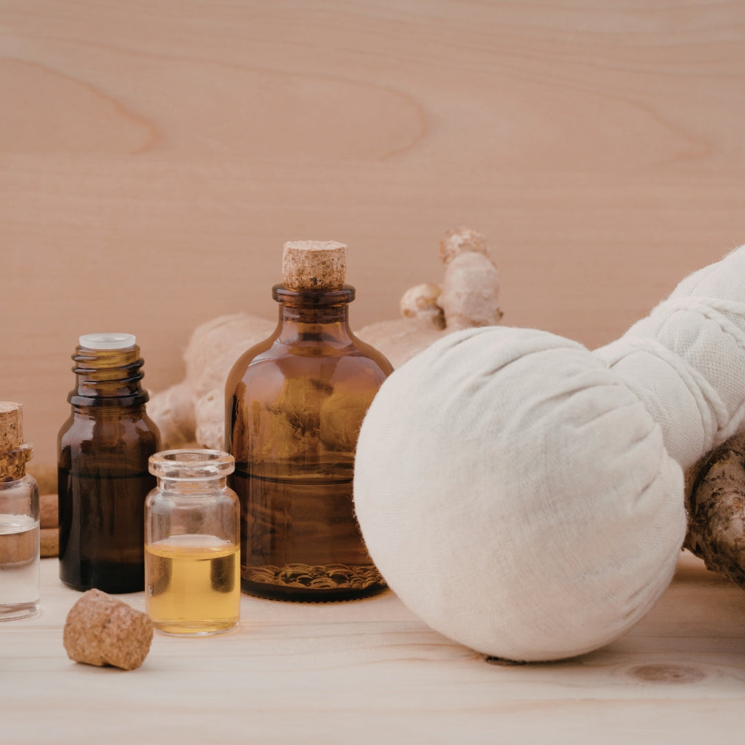 A large white massage ball rests on a light wood surface alongside amber glass bottles, small jars, and ginger roots against a matching wood background.