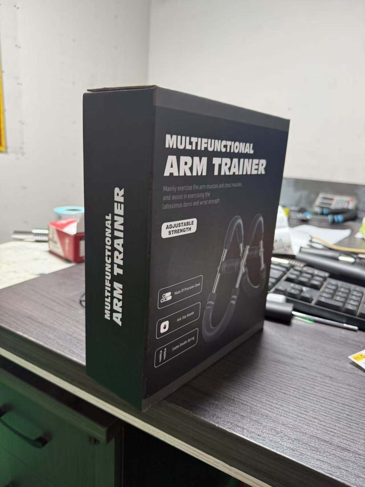 Multi-functional U-shaped Arm Strengthener in black packaging with adjustable strength and workout icons, displayed on a desk