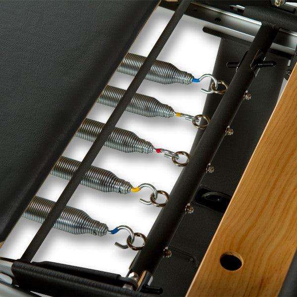 Close-up of black metal frame with coiled silver springs featuring colored hooks on wooden side panel, part of Peak PilateSystem® Deluxe Bundle equipment.