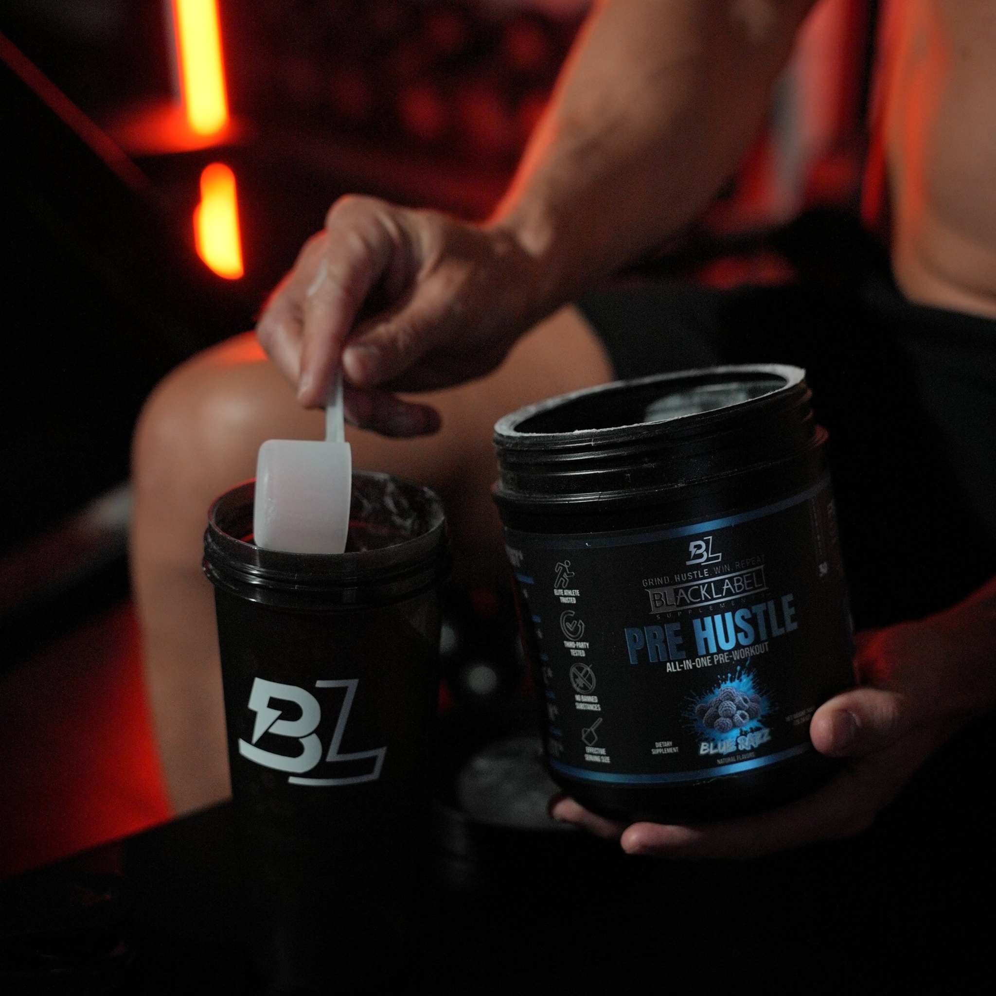PRE HUSTLE Pre-Workout black plastic tub with blue text and blueberry flavor label held by a person scooping powder into a black shaker cup with white logo