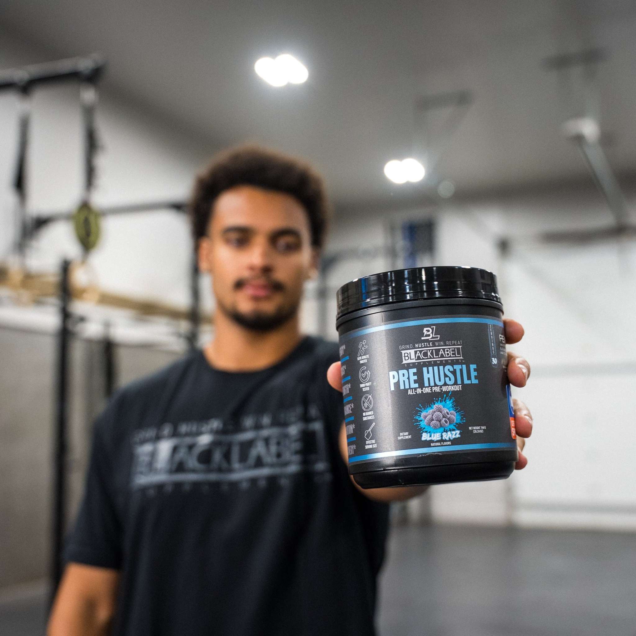Black plastic container of PRE HUSTLE Pre-Workout powder with blue and white label held by a man in a gym setting