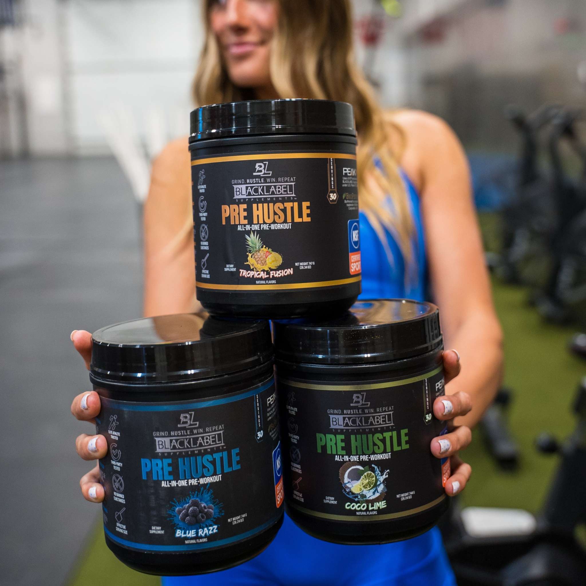 Three jars of PRE HUSTLE Pre-Workout in tropical fusion, blue razz, and coco lime flavors held by a woman in a blue tank top