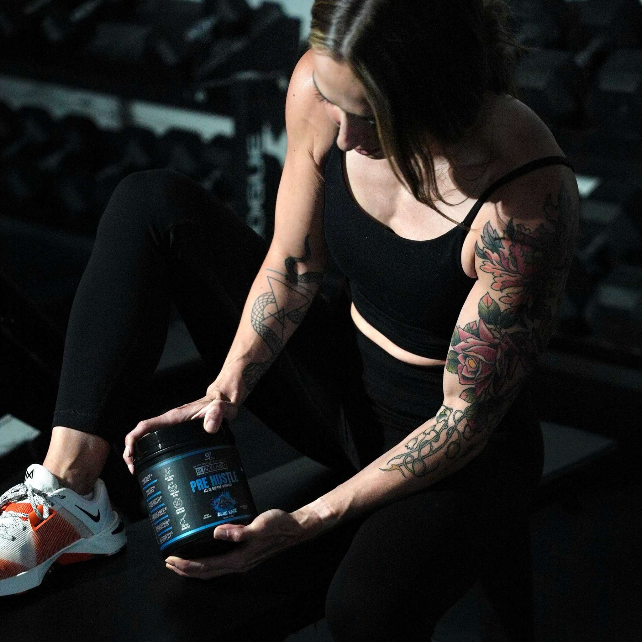 Black container of PRE HUSTLE Pre-Workout held by tattooed woman in black athletic wear with orange and white sneakers