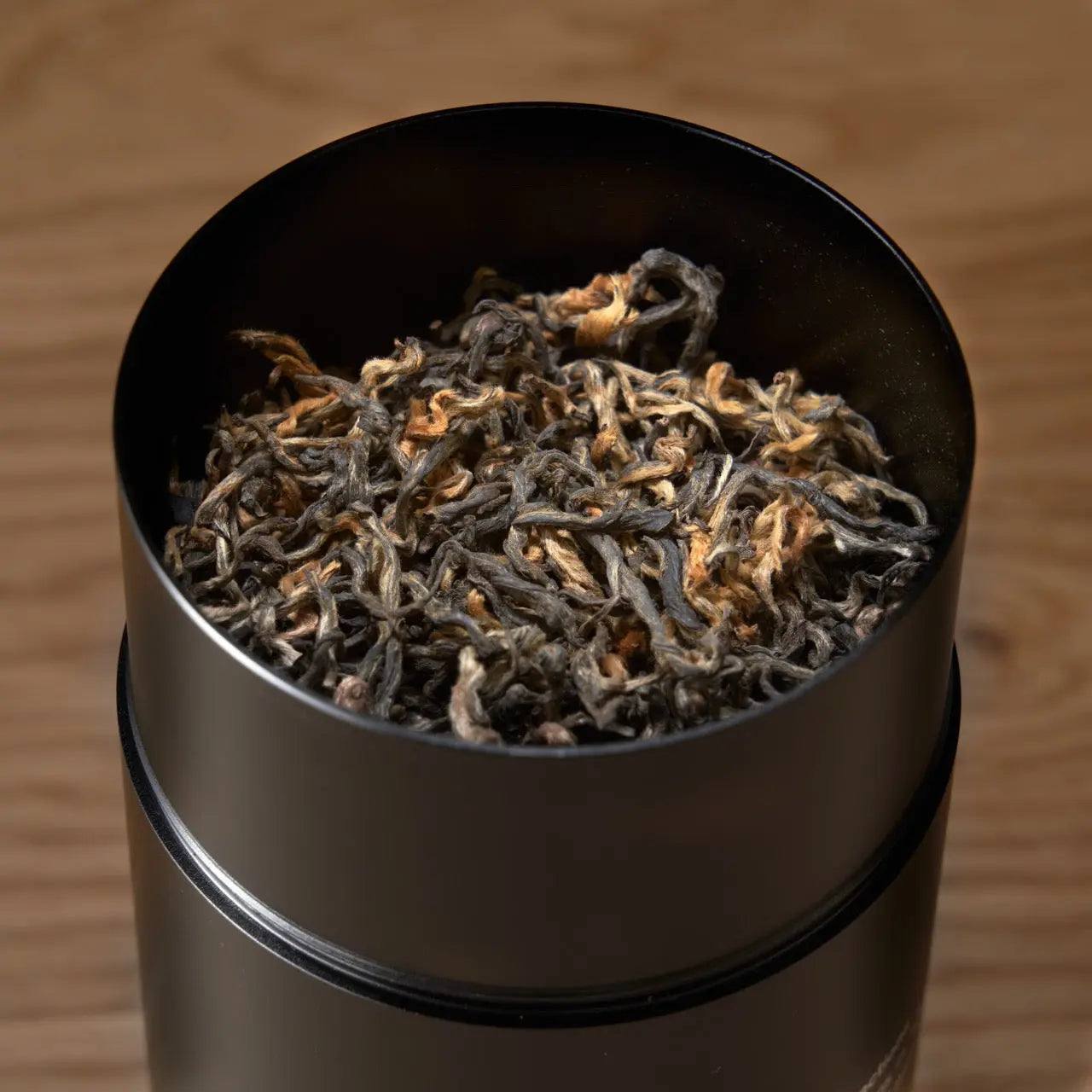 Premium Loose Leaf Tea in a black metal tin, filled with curled, textured brown and silver tea leaves