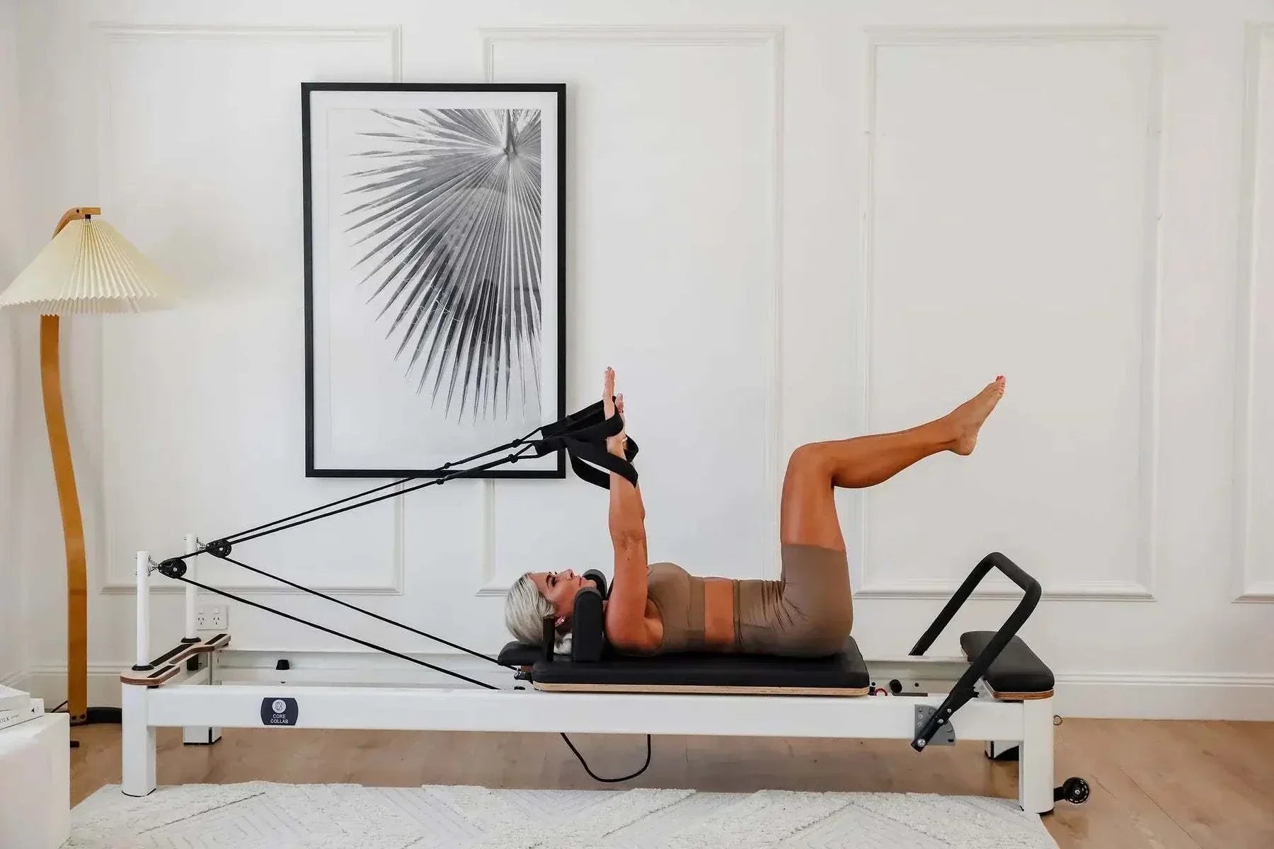 Queen Studio Pilates Reformer with white metal frame, black padded carriage, adjustable ropes, and footbar in use by a woman exercising.