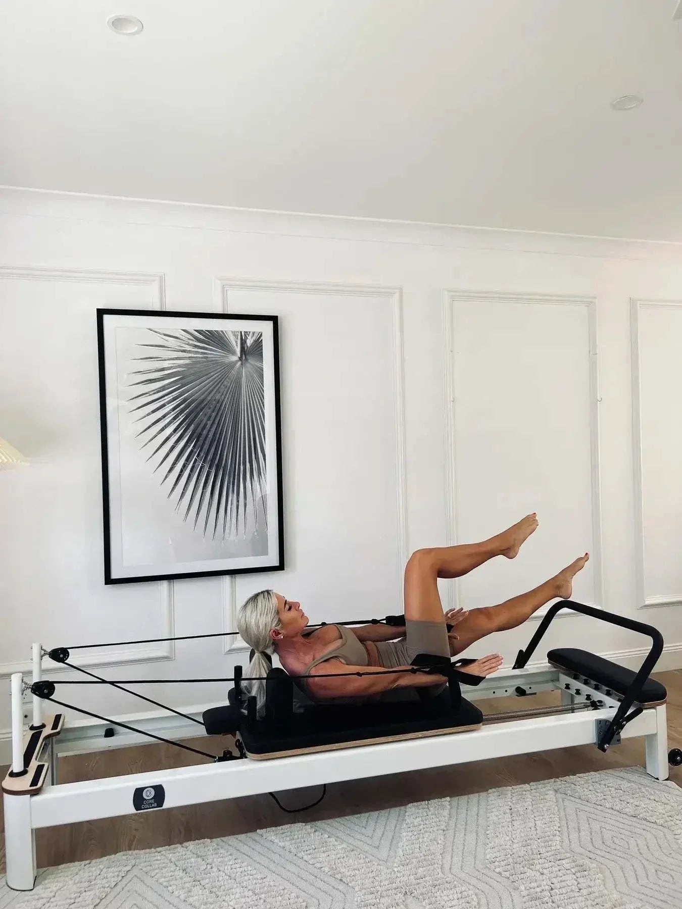 Queen Studio Pilates Reformer with white frame, black padded carriage, adjustable ropes and footbar, in use by a person performing leg exercises.
