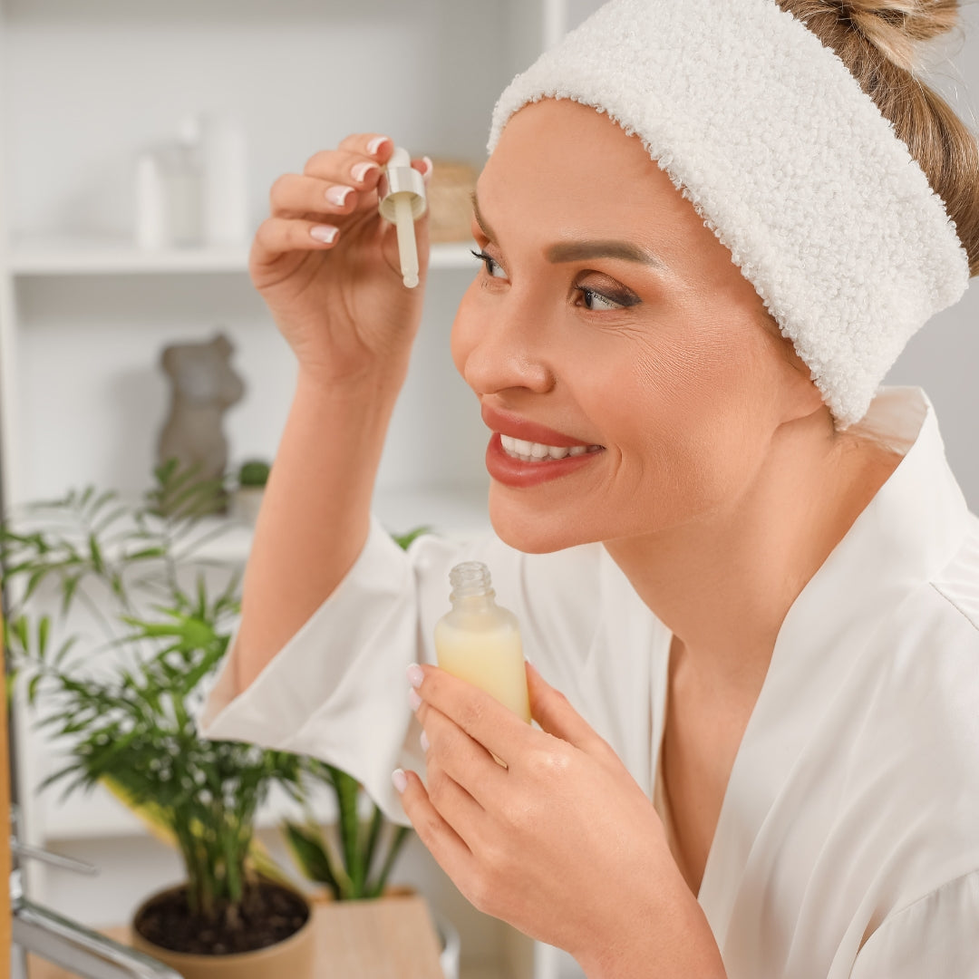 A smiling woman with a white textured headband and white robe applies a skincare serum from a dropper bottle to her face in a bright bathroom setting.