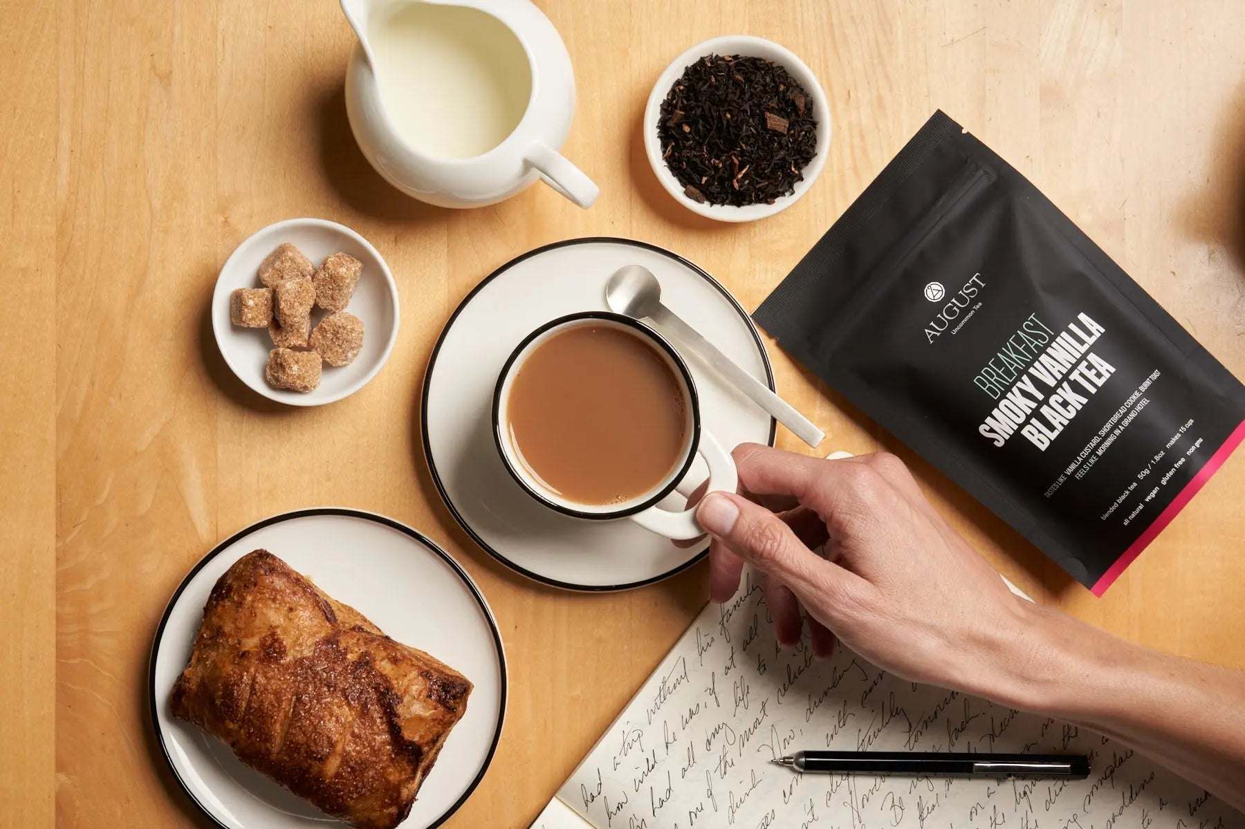 Smoky Vanilla Black Tea in a black pouch beside brewed tea, loose leaves, milk, sugar cubes and toast