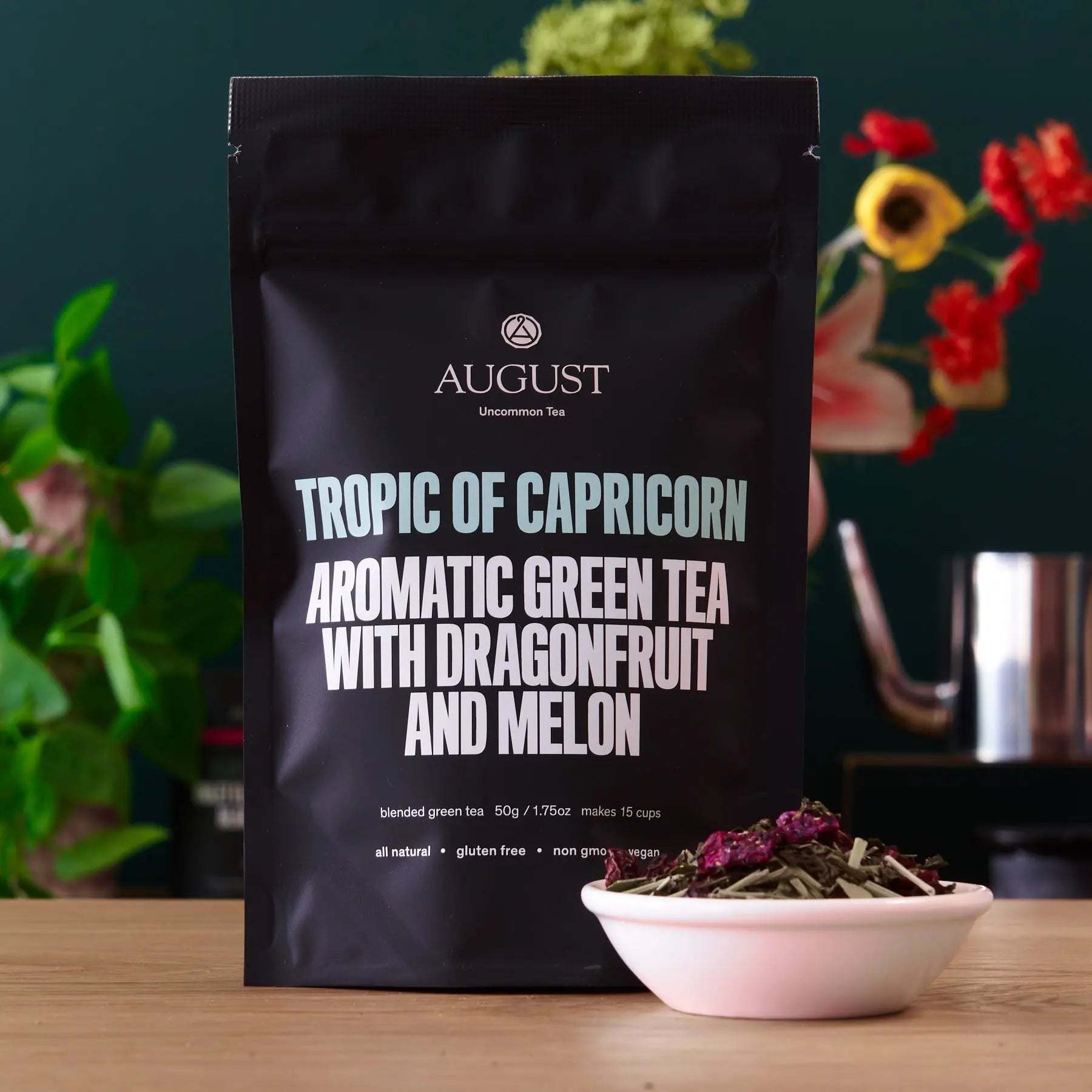 Tropic of Capricorn green tea in a matte black pouch with bold white lettering, beside loose tea in a pink bowl